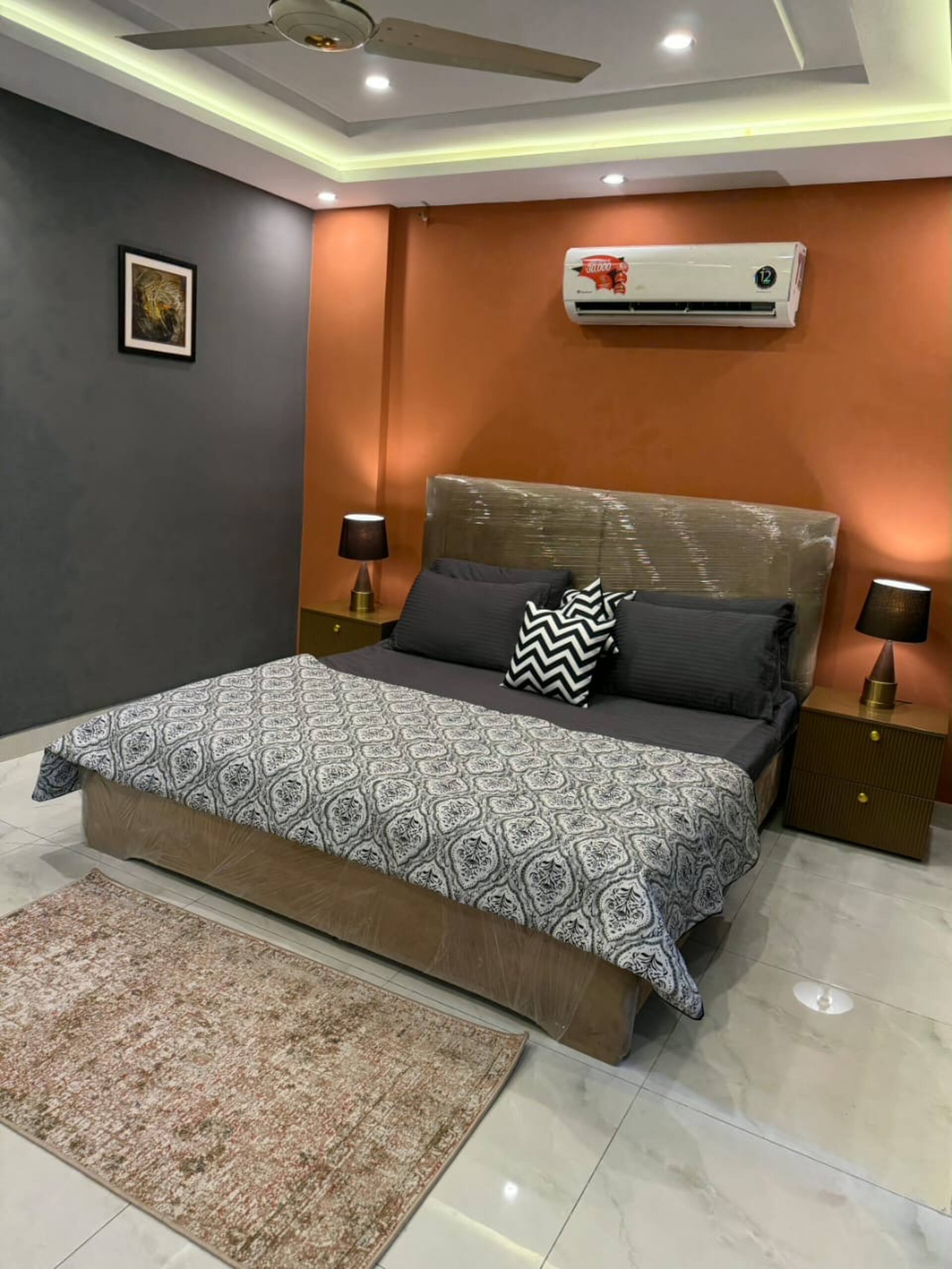 King bed with patterned linen and amber wall lighting in The Amber Haven, the Aangan one-bedroom apartment facing Grand Jamia Mosque in Bahria Town Lahore