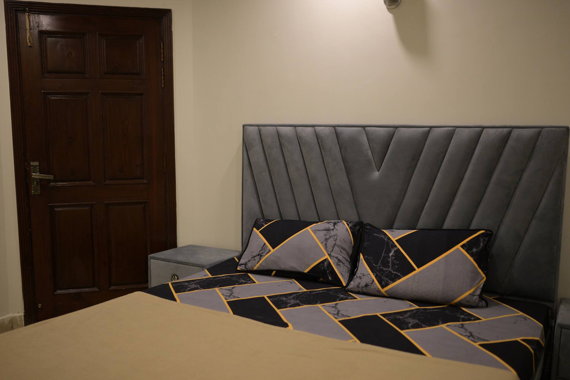 Double bed with a grey upholstered headboard and patterned bedding in the Aangan three-bedroom gaming suite in Apollo Tower, E-11 Islamabad