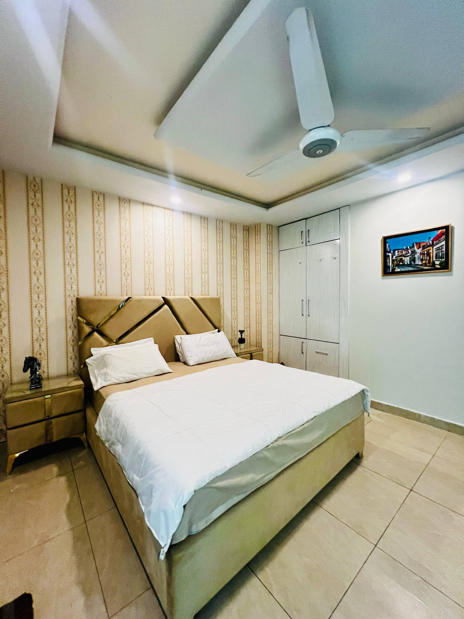 Third bedroom with a double bed, ceiling fan and wall-mounted TV in the Aangan three-bedroom gaming suite in Apollo Tower, E-11 Islamabad
